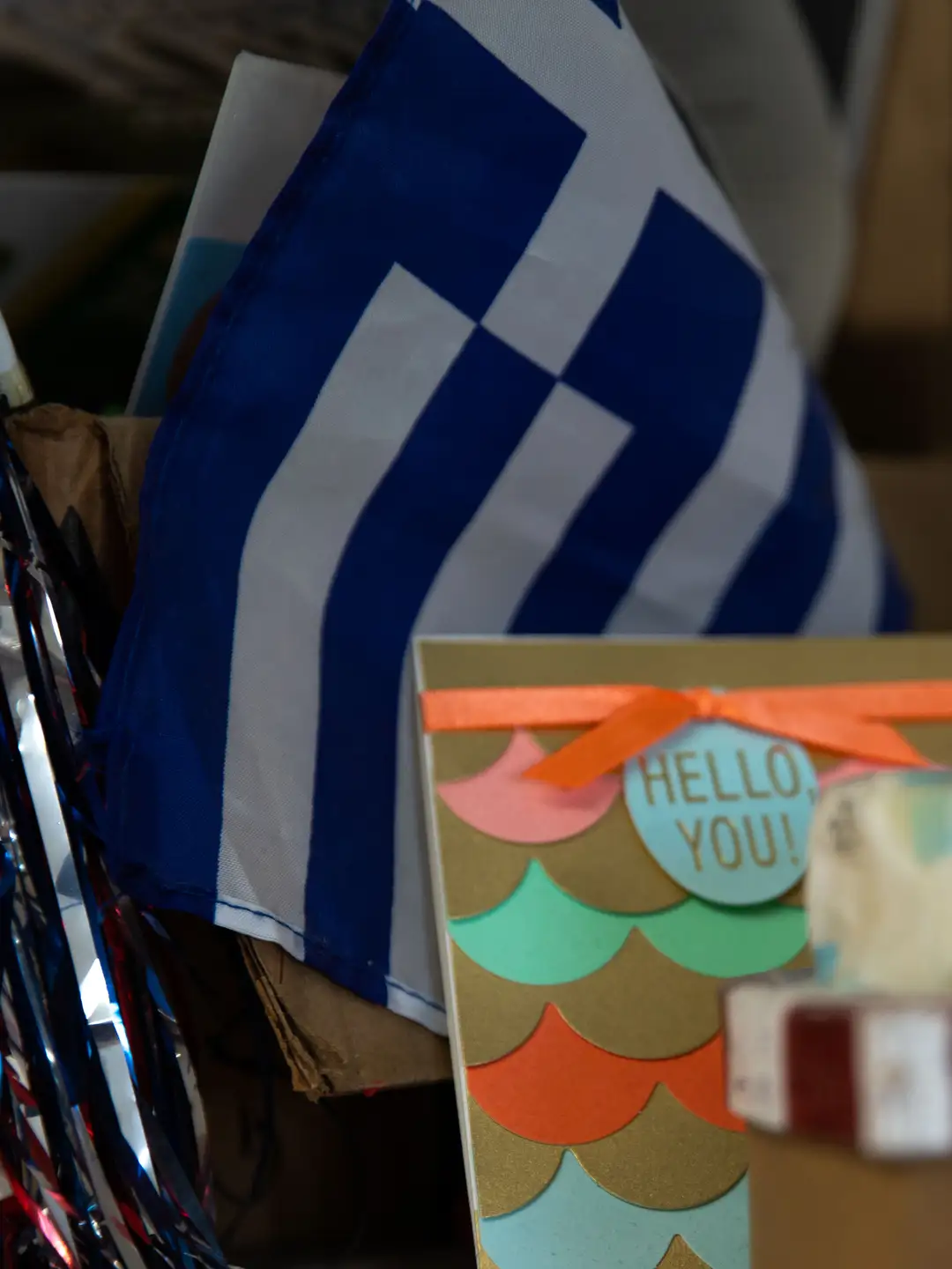 Κάρτα με την ένδειξη «HELLO, YOU!», ανάμεσα σε τυλιγμένα αντικείμενα και μια ελληνική σημαία.