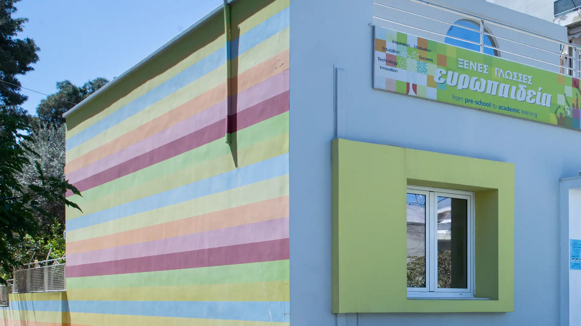 Η πρόσοψη του Green School: χρωματιστές οριζόντιες ρίγες, κόκκινη πόρτα και η πινακίδα «ξένες γλώσσες ευρωπαιδεία».
