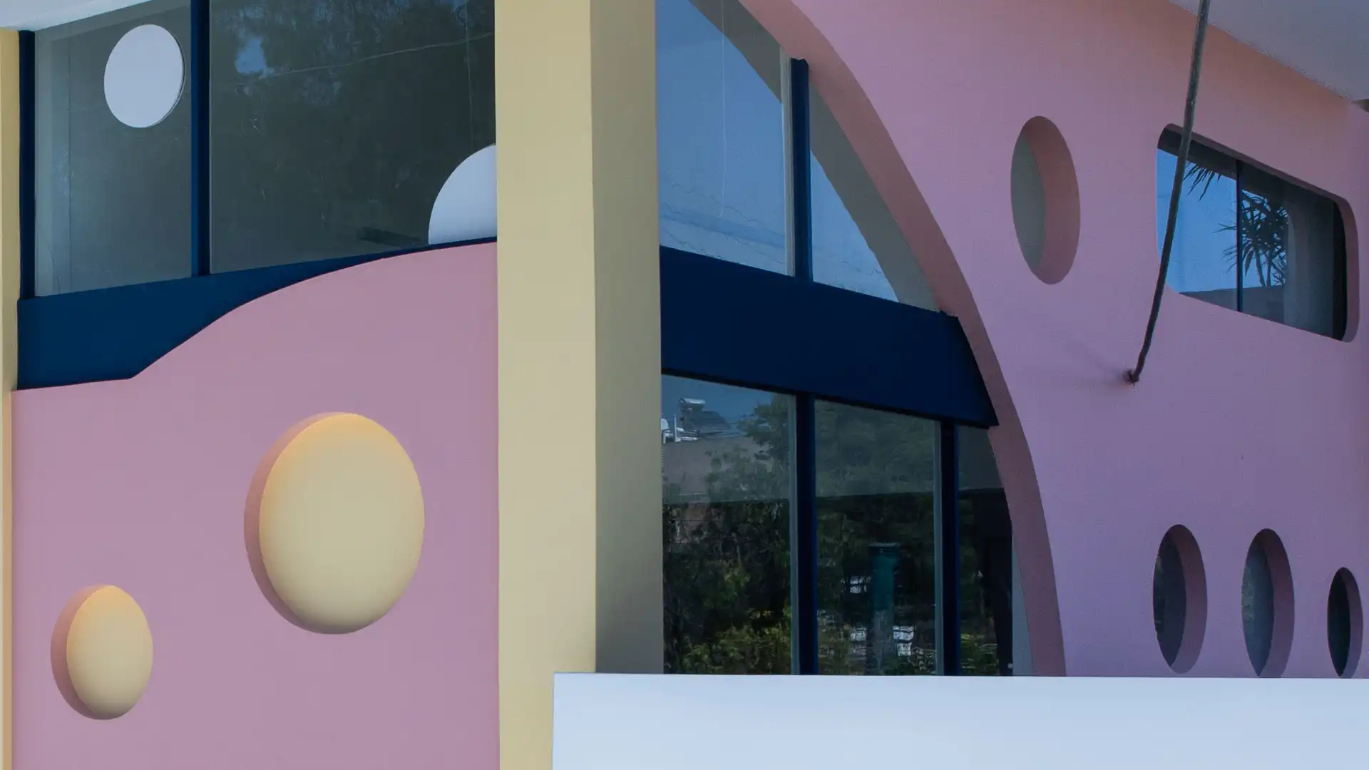 Η πρόσοψη του New Generation School: μπλε πινακίδα «ξένες γλώσσες ευρωπαιδεία», ροζ τοίχος με στρογγυλά ανοίγματα και ζωγραφισμένος αύλειος χώρος με παγκάκια.
