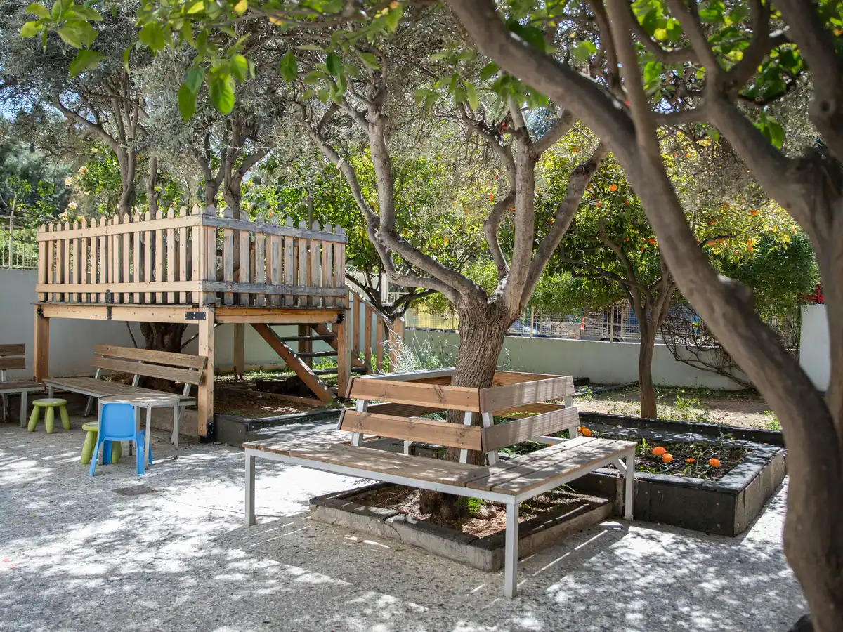 Ο κήπος του Green School: ξύλινο πατάρι με κάγκελα, ελιά και πορτοκαλιές, παγκάκια, χαμηλά παρτέρια με φυτά και μικρά σκαμπό.