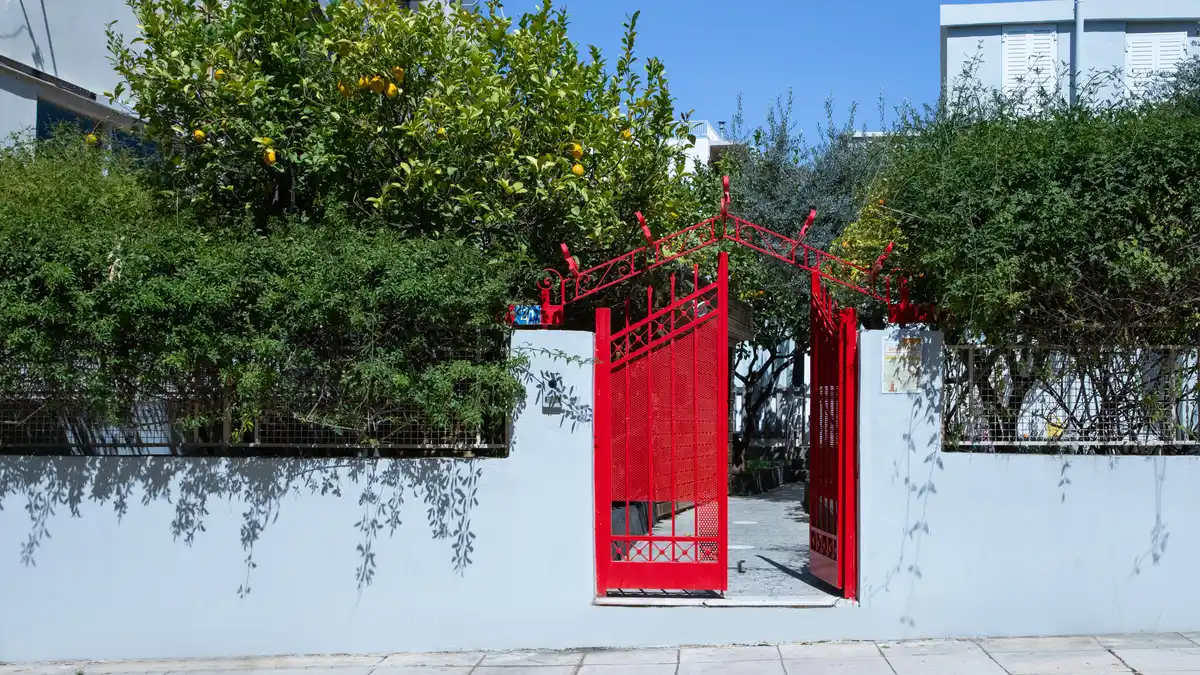 Η είσοδος του Green School: κόκκινη σιδερένια καγκελόπορτα σε λευκό τοίχο, με μια λεμονιά και πυκνή φυλλωσιά από πάνω, και πλακόστρωτο πεζοδρόμιο μπροστά.