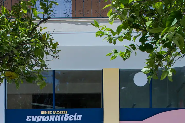 Η πρόσοψη σχολείου της Ευρωπαιδείας: η μπλε πινακίδα «ΞΕΝΕΣ ΓΛΩΣΣΕΣ ευρωπαιδεία» κάτω από το καμπύλο λευκό γείσο, με φυλλωσιά λεμονιάς μπροστά και την άκρη του ροζ τοίχου δεξιά.