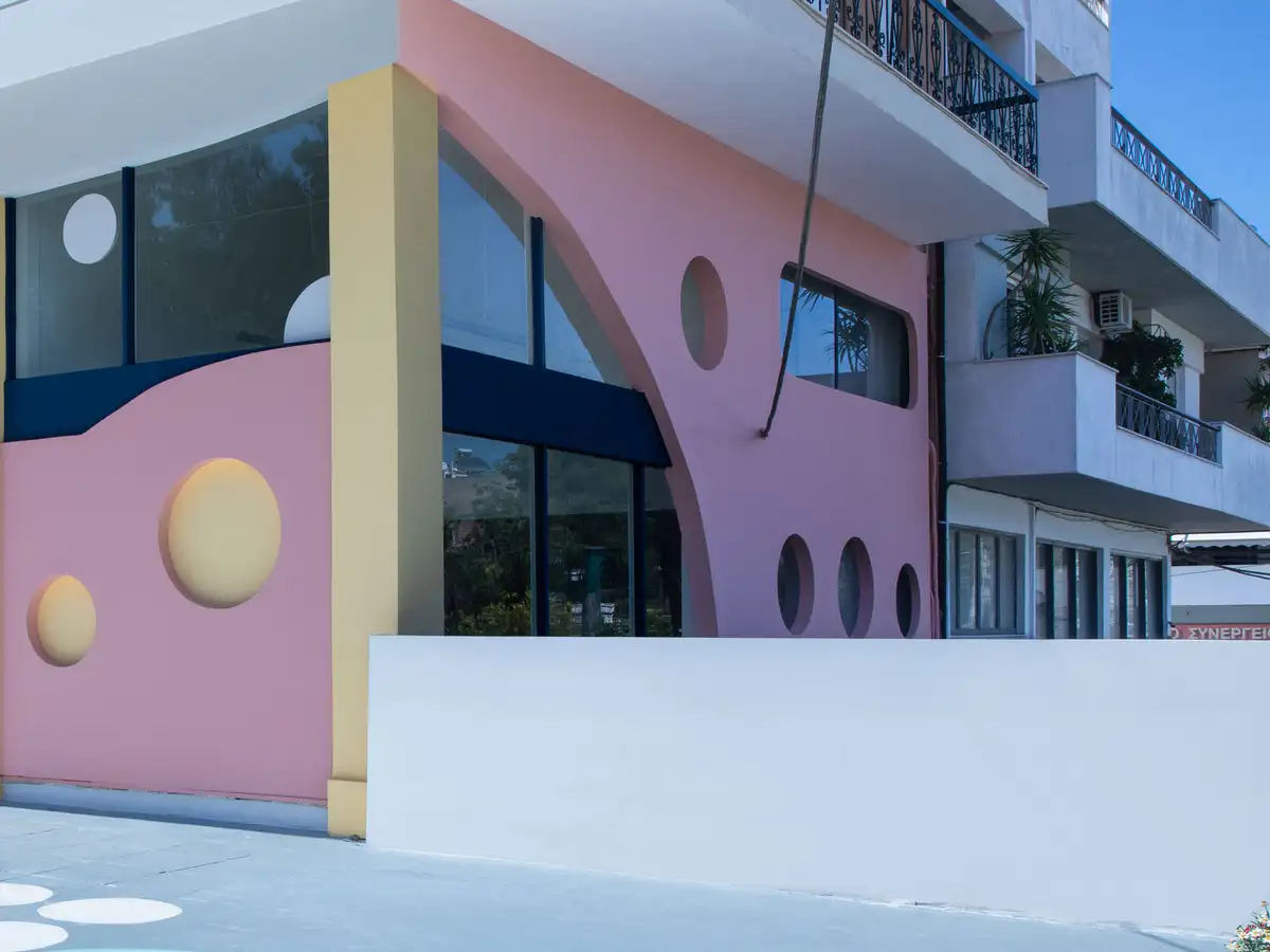Η γωνία του New Generation School: ροζ τοίχος με μια μεγάλη καμπύλη, στρογγυλά ανάγλυφα σε κίτρινο και στρογγυλά παράθυρα, κίτρινη κολόνα στη γωνία και χαμηλός λευκός τοίχος μπροστά.