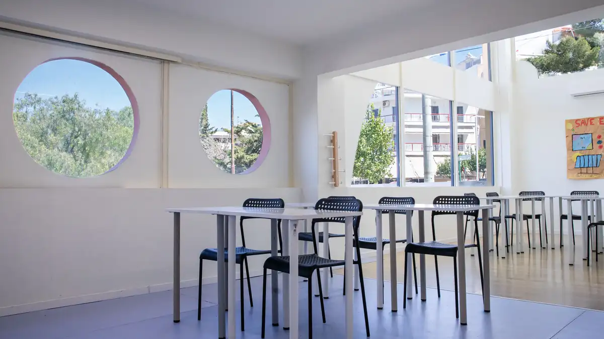 Αίθουσα του New Generation School: δύο στρογγυλά παράθυρα με ροζ πλαίσιο που βλέπουν σε δέντρα, λευκά θρανία με μαύρες καρέκλες, και μια αφίσα μαθητών στον απέναντι τοίχο.