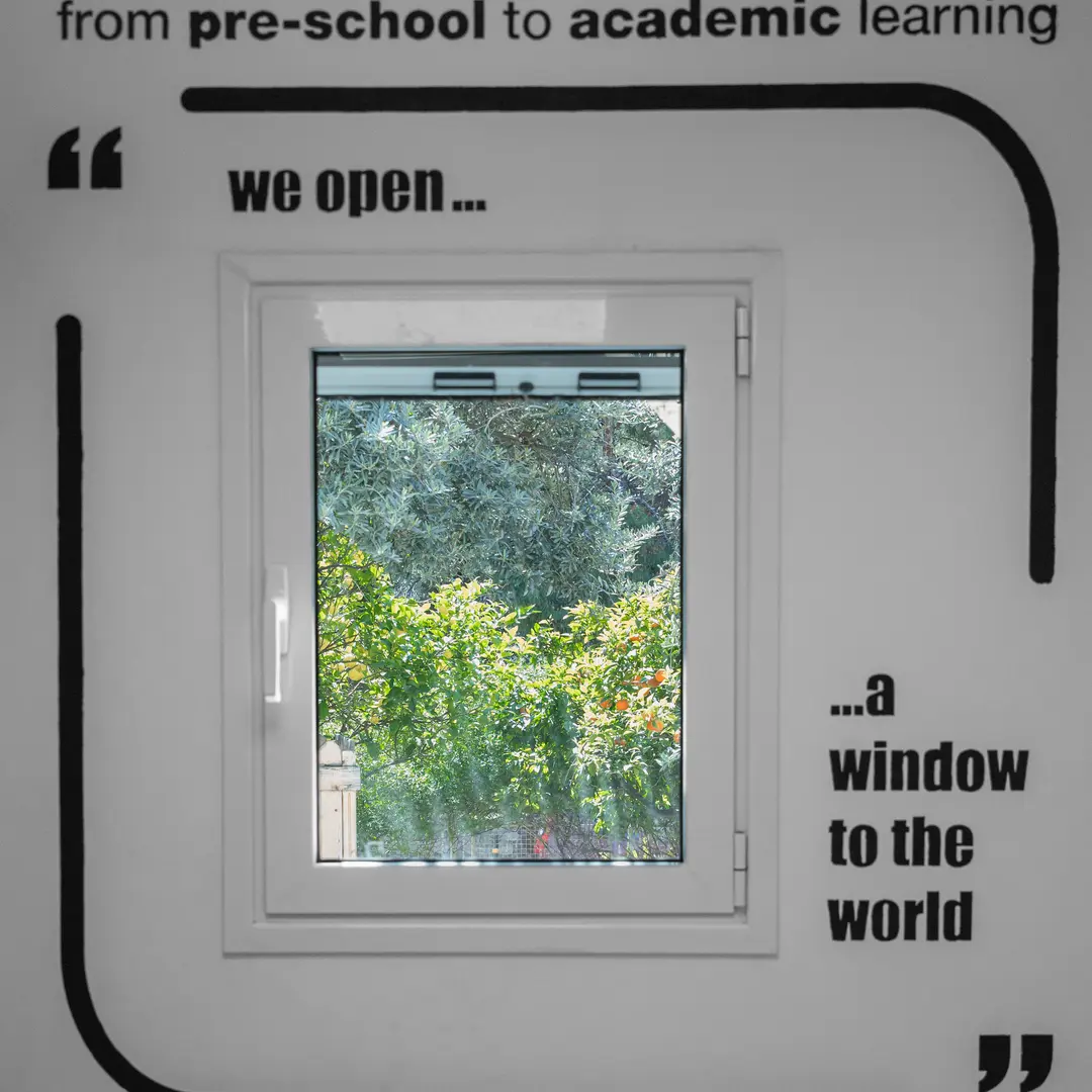 Τοίχος σχολείου με τη φράση «from pre-school to academic learning — we open a window to the world», γύρω από ένα παράθυρο που βλέπει στον κήπο.